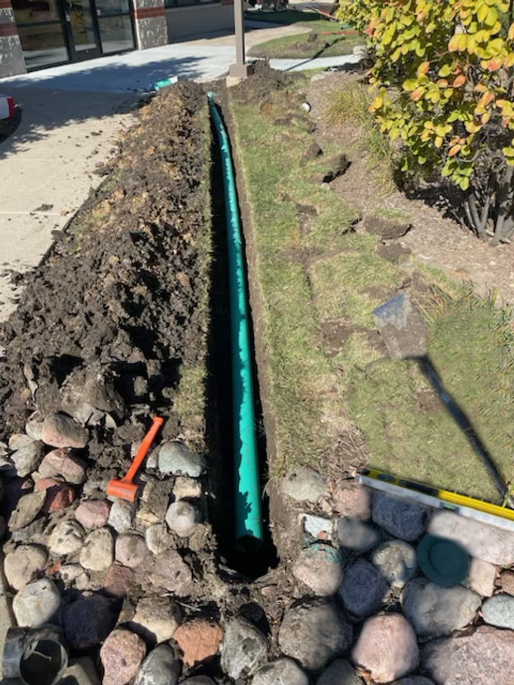 Trenched area with green drainage pipe, surrounded by soil and landscaping rocks.