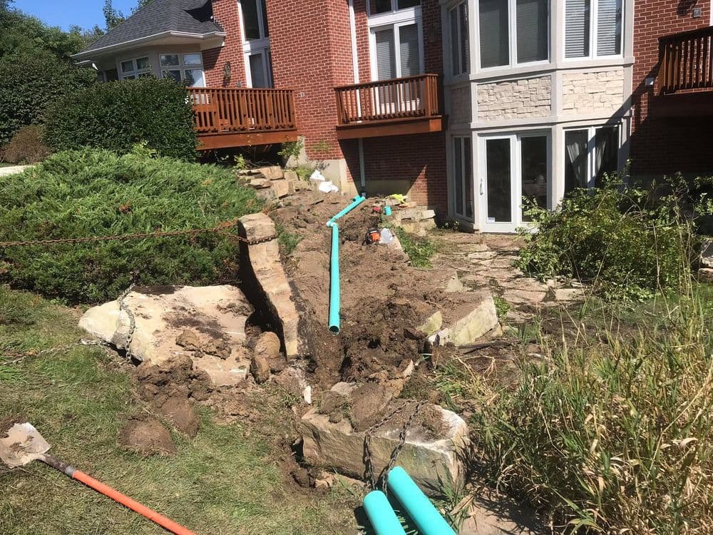 Construction site showing soil excavation and blue pipes near a residential backyard.