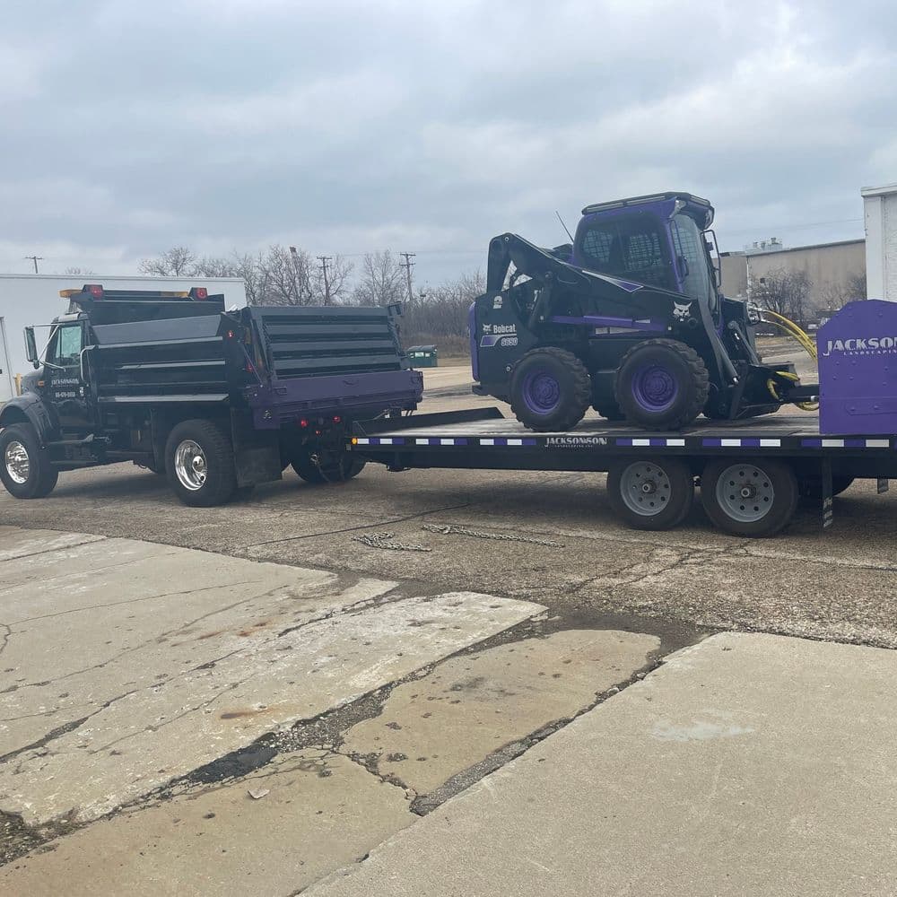 Dump truck towing a trailer with a compact track loader for construction work.